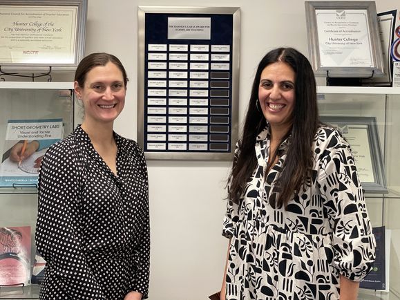 Kathryn Ahmed and Nicora Placa stand in front of the Ladas Award plaque displaying their names.